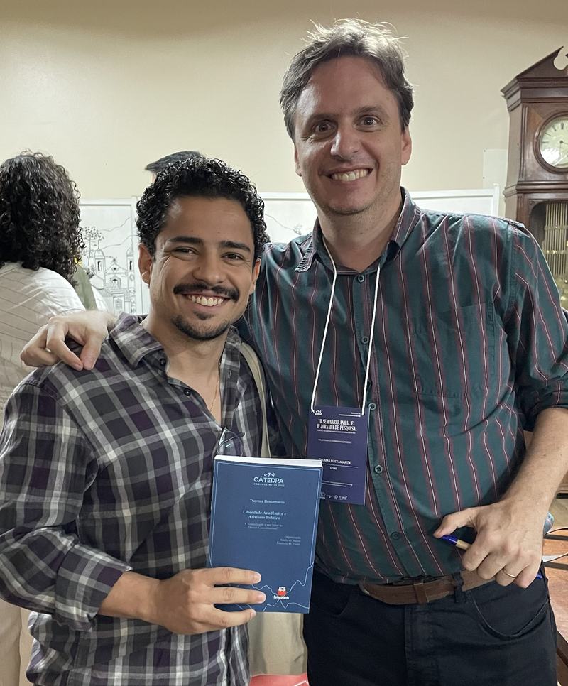 Victor segurando um livro azul, abraçado a Thomas Bustamante
