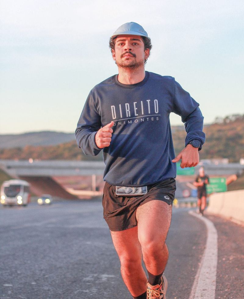 Victor correndo numa rodovia ao amanhecer, de boné e moletom do Direito da Unimontes