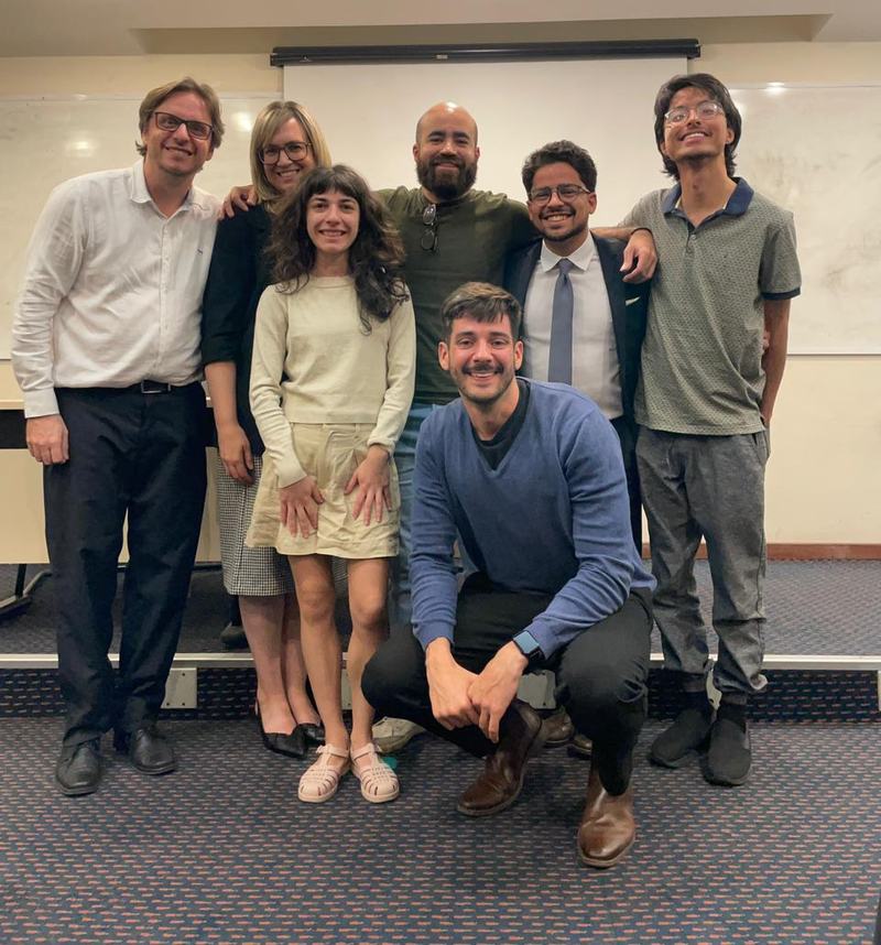 Grupo de sete pessoas posando diante de um quadro branco numa sala de aula