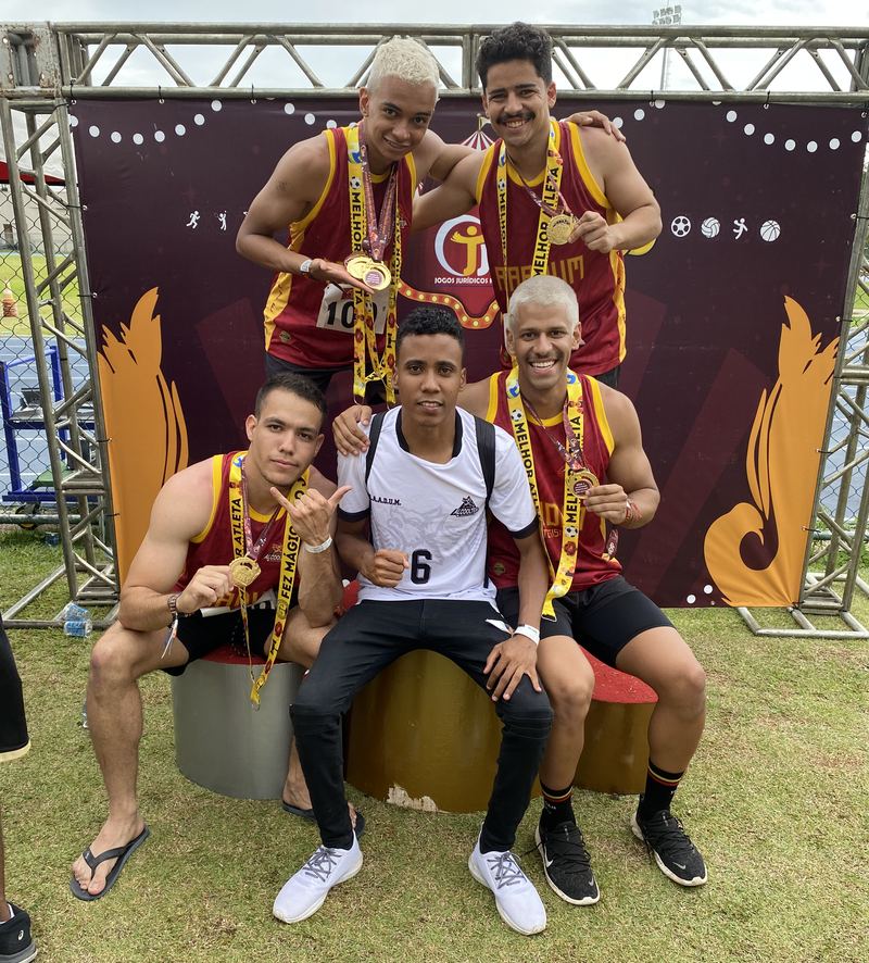 Equipe de atletismo com medalhas de ouro no pescoço, posando diante de um painel dos jogos